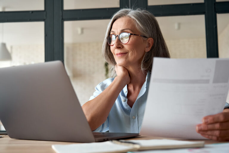 Lachende volwassen zakenvrouw van middelbare leeftijd die een laptop gebruikt en aan haar bureau werkt. Gelukkige oudere zakenvrouw (HR) houdt een cv vast en interviewt een sollicitant op afstand, of een oudere werkzoekende die online naar een baan zoekt.